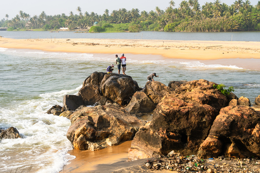 View of people standing on large coastal rocks with waves crashing against the shore and lush palm trees in the background Galle Southern Province Sri Lanka.