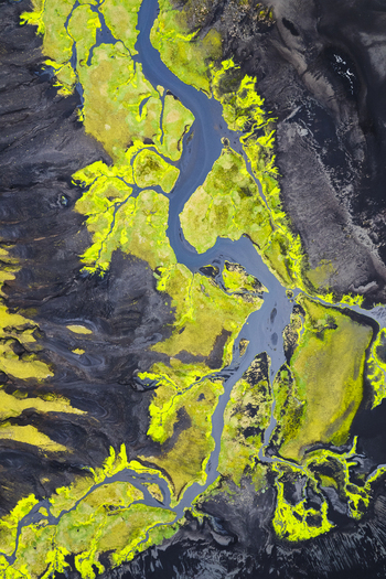 Aerial view of a winding blue glacial river cutting through vibrant green moss and dark volcanic soil near the F232 mountain road in Skaftarhreppur Iceland.
