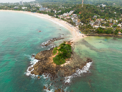 Aerial view of Parrot Rock island connected to the sandy beach by a narrow sandbar with turquoise ocean water and coastal buildings Mirissa Southern Province Sri Lanka. Aerial view of Parrot Rock island connected to the sandy beach by a narrow sandbar with turquoise ocean water and coastal buildings Mirissa Southern Province Sri Lanka.