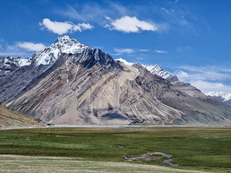 View of Penzi La road winding through the lush green valley with a small stream and snow capped mountains in the background under a clear blue sky in Zanskar Ladakh India.