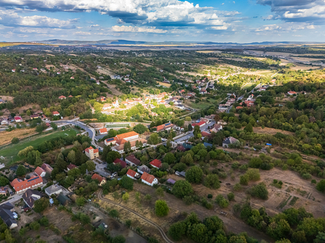 Aerial view of Csabdi village in Hungary with red roofed homes a church winding roads and small farms set among wooded hills fields and a broad rural valley.