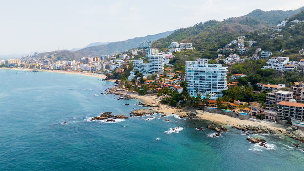 Aerial view of turquoise waters embrace the rugged coastline where modern buildings meet traditional homes amidst lush green hills Puerto Vallarta Jalisco Mexico.