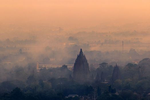 Aerial view of Prambanan Temple rising through the morning mist surrounded by lush tropical trees and historic structures in Kecamatan Prambanan Daerah Istimewa Yogya Indonesia.