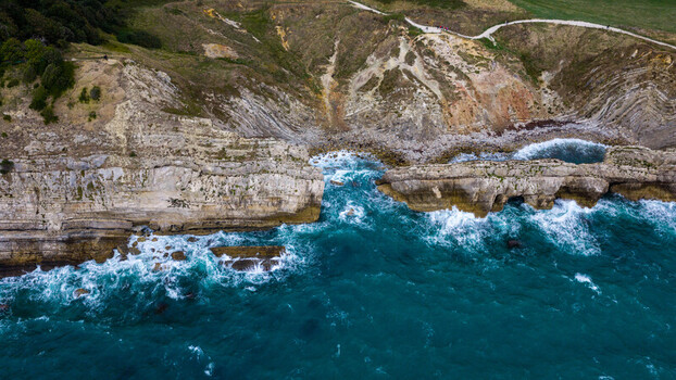 Aerial view of Lulworth Cove featuring dramatic limestone cliffs turquoise sea crashing waves and a coastal path winding along the rugged terrain in Wareham England United Kingdom. Aerial view of Lulworth Cove featuring dramatic limestone cliffs turquoise sea crashing waves and a coastal path winding along the rugged terrain in Wareham England United Kingdom.