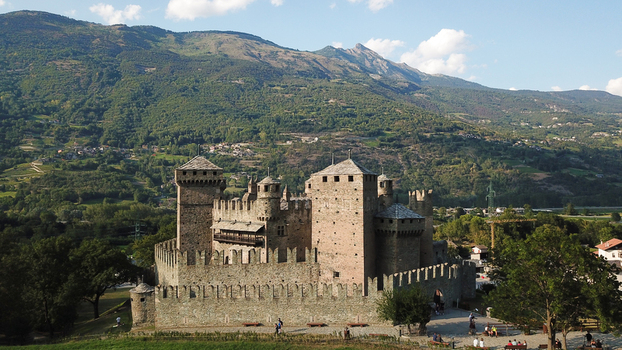 Aerial view of Fenis Castle a medieval stone fortress with people walking near the entrance set against the lush green mountains of the Alps Aosta Valley Aosta Valley Italy. Aerial view of Fenis Castle a medieval stone fortress with people walking near the entrance set against the lush green mountains of the Alps Aosta Valley Aosta Valley Italy.