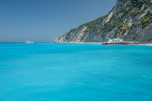 View of Egremni Beach with turquoise water a large excursion boat docked at the shore and steep white cliffs under a clear blue sky in Lefkada Greece.