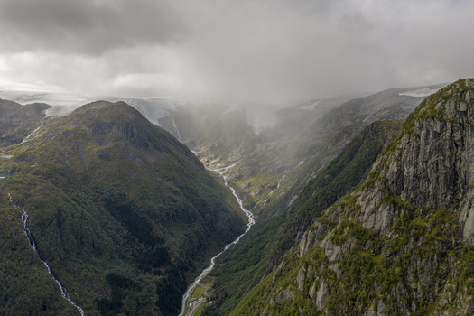 Aerial view of the Buarbreen glacier and a winding river flowing through a deep mountain valley under a misty sky in Ullensvang Vestland Norway. Aerial view of the Buarbreen glacier and a winding river flowing through a deep mountain valley under a misty sky in Ullensvang Vestland Norway.