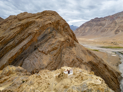 Aerial view of a remote Buddhist monastery perched on a rocky cliffside overlooking a winding river valley in the rugged Himalayan landscape of Zanskar Ladakh India.