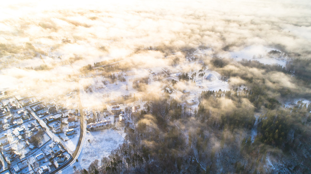 Aerial view of a snow covered village nestled among dense forests and rolling hills partially obscured by low hanging morning mist illuminated by golden sunlight in Latvia. Aerial view of a snow covered village nestled among dense forests and rolling hills partially obscured by low hanging morning mist illuminated by golden sunlight in Latvia.