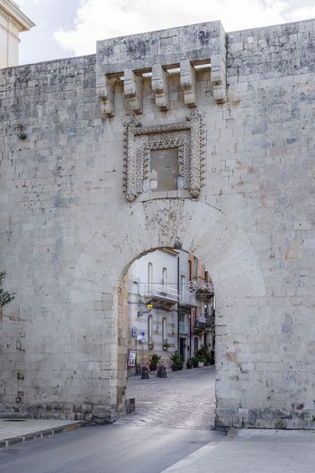 View of the ancient stone archway leading into a historic street casting shadows on the cobblestone Ortigia Sicily Italy.