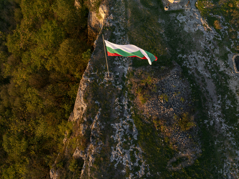 Aerial view of the Bulgarian flag waving on a pole atop the rocky cliffs of Ovech Fortress surrounded by lush forest and rugged terrain at sunset in Provadia Varna Bulgaria. Aerial view of the Bulgarian flag waving on a pole atop the rocky cliffs of Ovech Fortress surrounded by lush forest and rugged terrain at sunset in Provadia Varna Bulgaria.