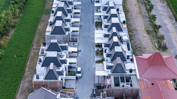 Aerial view of a modern residential street with uniform houses featuring gray roofs and white walls flanked by green fields and construction areas in Indonesia. Aerial view of a modern residential street with uniform houses featuring gray roofs and white walls flanked by green fields and construction areas in Indonesia.