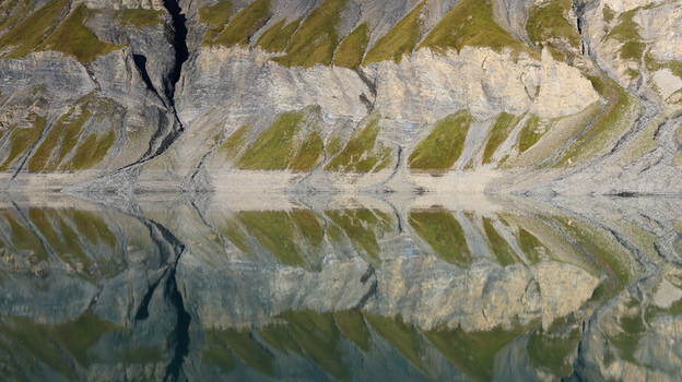 View of the calm waters of Lac du Vieux Emosson reflecting the steep rocky mountain slopes with patches of green grass in Finhaut Valais Switzerland. View of the calm waters of Lac du Vieux Emosson reflecting the steep rocky mountain slopes with patches of green grass in Finhaut Valais Switzerland.