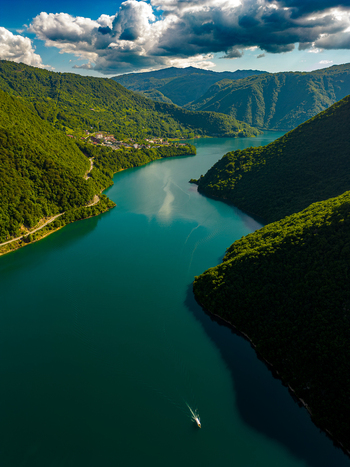 Aerial view of Piva Lake with a small boat navigating the turquoise waters surrounded by lush green mountains under a dramatic cloudy sky in Pluzine Municipality Montenegro. Aerial view of Piva Lake with a small boat navigating the turquoise waters surrounded by lush green mountains under a dramatic cloudy sky in Pluzine Municipality Montenegro.