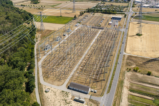 Aerial view of the Amprion Umspannwerk electrical substation with metal infrastructure power lines and transformers surrounded by rural fields and forest in Pfungstadt Hessen Germany.