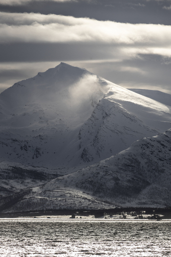 View of snow covered mountain peaks and a fjord under a dramatic cloudy sky with a small coastal settlement at the base in Tromso Troms Norway.