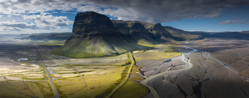 Aerial view of the massive Lomagnupur mountain rising above green plains and a braided glacial river system under a dramatic cloudy sky in Iceland.