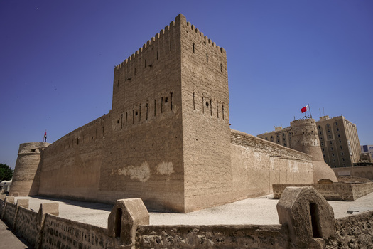 View of the historic Al Fahidi Fort with its traditional sand colored stone walls defensive towers and a waving flag under a clear blue sky in Dubai Dubai United Arab Emirates. View of the historic Al Fahidi Fort with its traditional sand colored stone walls defensive towers and a waving flag under a clear blue sky in Dubai Dubai United Arab Emirates.