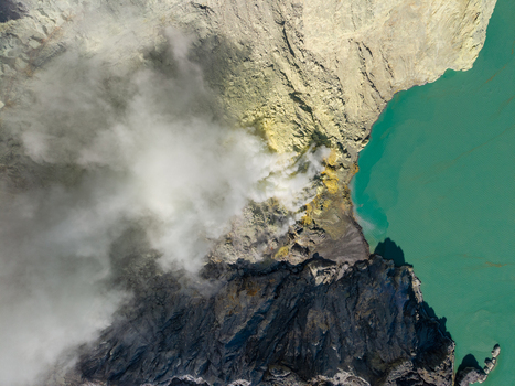Aerial view of the turquoise crater lake and volcanic sulfur mining area at Mount Ijen with thick plumes of smoke rising from the rugged terrain in Ijen East Java Indonesia.