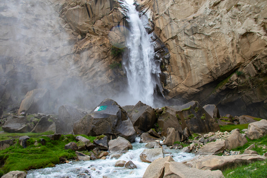 View of Khamosh Waterfall cascading down steep rocky cliffs into a flowing stream with large boulders and green grass in Skardu Gilgit Pakistan.
