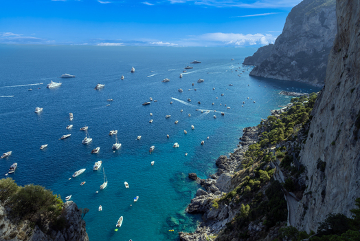 Aerial view of Capri with numerous boats and yachts anchored in the turquoise bay surrounded by dramatic steep cliffs and lush greenery in Capri Campania Italy. Aerial view of Capri with numerous boats and yachts anchored in the turquoise bay surrounded by dramatic steep cliffs and lush greenery in Capri Campania Italy.
