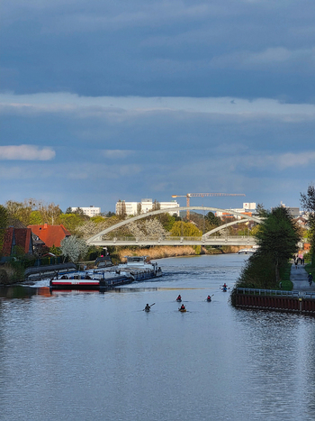 Hannover Germany 14 April 2026 View of a cargo barge on the canal under a modern arch bridge with kayakers paddling surrounded by spring trees and urban buildings under a cloudy sky.