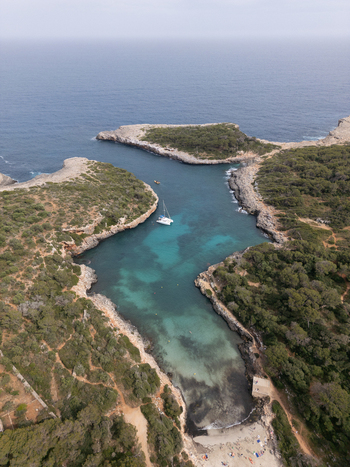 Aerial view of Cala Sa Nau featuring turquoise waters rocky cliffs with green trees a white catamaran and people on the beach in Felanitx Balearic Islands Spain. Aerial view of Cala Sa Nau featuring turquoise waters rocky cliffs with green trees a white catamaran and people on the beach in Felanitx Balearic Islands Spain.