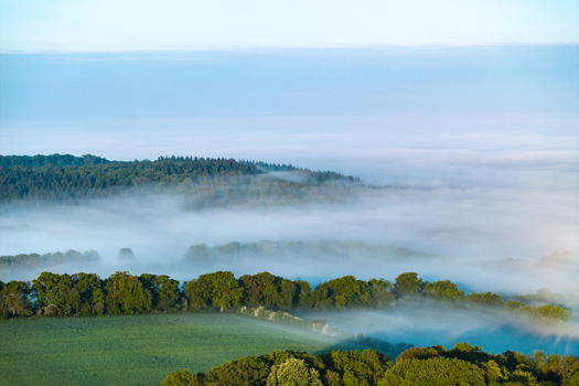 Aerial view of rolling hills and lush green forests partially covered by thick morning mist under a clear blue sky in Nuffield Oxfordshire United Kingdom. Aerial view of rolling hills and lush green forests partially covered by thick morning mist under a clear blue sky in Nuffield Oxfordshire United Kingdom.