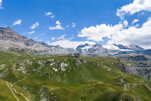 Aerial view of Breuil Cervinia nestled in the green slopes of the Aosta Valley with snow capped Alps and a glacier in the background under a blue sky Breuil Cervinia Aosta Valley Italy. Aerial view of Breuil Cervinia nestled in the green slopes of the Aosta Valley with snow capped Alps and a glacier in the background under a blue sky Breuil Cervinia Aosta Valley Italy.