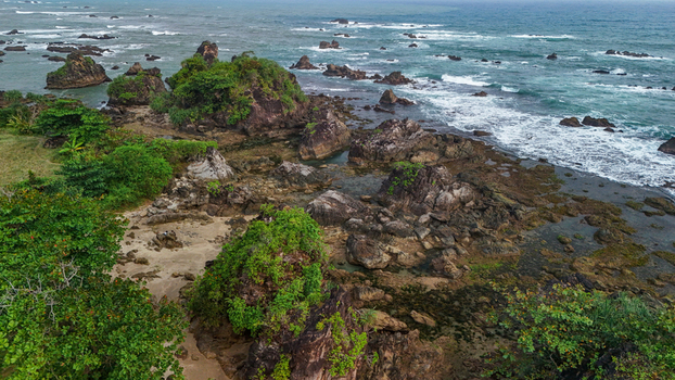 Aerial view of a rugged coastline featuring dark volcanic rock formations lush green vegetation and waves crashing against the shore in Indonesia. Aerial view of a rugged coastline featuring dark volcanic rock formations lush green vegetation and waves crashing against the shore in Indonesia.