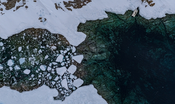 Aerial view of a small boat moored by a snow covered rocky shoreline with turquoise water and floating ice floes in Tasiilaq Sermersooq Municipality Greenland. Aerial view of a small boat moored by a snow covered rocky shoreline with turquoise water and floating ice floes in Tasiilaq Sermersooq Municipality Greenland.