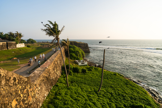 View of the historic Galle Fort ramparts with people walking along the stone walls leaning palm trees and the Indian Ocean coastline in Galle Southern Province Sri Lanka.