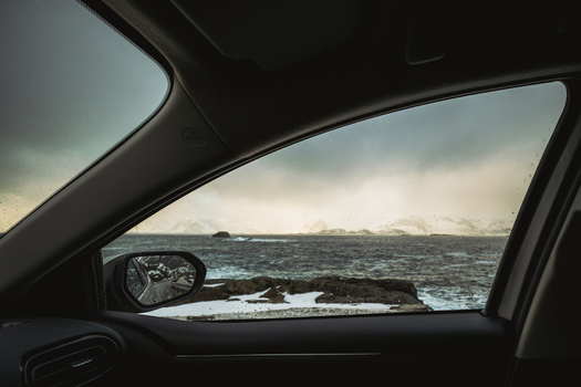 View of choppy ocean waters meet rugged snow dusted islands under a cloudy sky framed by a car window s dark interior Henningsv&aelig;r Nordland Norway.