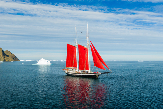 Aerial view of a sailboat with vibrant red sails navigating through Arctic waters surrounded by icebergs under a clear blue sky in Greenland. Aerial view of a sailboat with vibrant red sails navigating through Arctic waters surrounded by icebergs under a clear blue sky in Greenland.