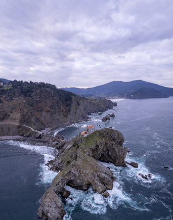 Aerial view of San Juan de Gaztelugatxe on a rocky islet connected by a stone bridge surrounded by the Cantabrian Sea cliffs and cloudy sky in Gaztelugatxeko Doniene Euskadi Spain.