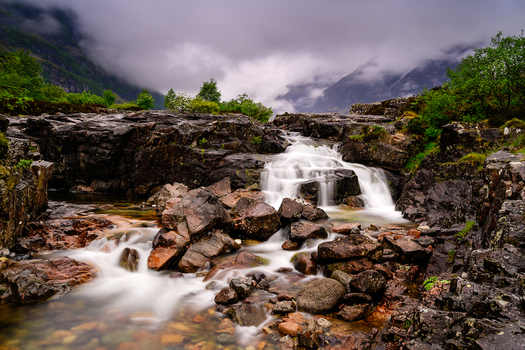 View of a cascading waterfall flowing over dark mossy rocks in a rugged landscape under a cloudy sky in Isle of Skye Scotland. View of a cascading waterfall flowing over dark mossy rocks in a rugged landscape under a cloudy sky in Isle of Skye Scotland.