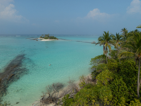 Aerial view of pristine turquoise waters gently lapping a palm fringed shore with a distant sandy islet connected by a shallow reef Thuraakunu Thiladhunmathee Uthuruburi Maldives. Aerial view of pristine turquoise waters gently lapping a palm fringed shore with a distant sandy islet connected by a shallow reef Thuraakunu Thiladhunmathee Uthuruburi Maldives.