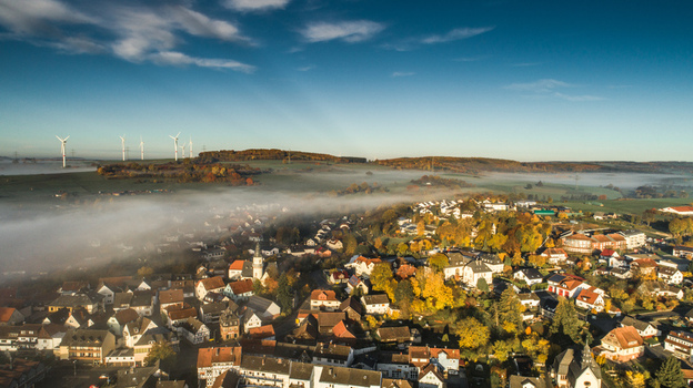 Aerial view of a German village nestled in a valley during autumn with colorful trees morning mist and wind turbines on the horizon in Germany. Aerial view of a German village nestled in a valley during autumn with colorful trees morning mist and wind turbines on the horizon in Germany.