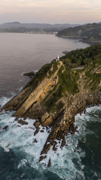 Aerial view of Getariako Itsasargia lighthouse perched on a dramatic rocky cliff surrounded by crashing waves and lush greenery at sunset in Getaria Euskadi Spain. Aerial view of Getariako Itsasargia lighthouse perched on a dramatic rocky cliff surrounded by crashing waves and lush greenery at sunset in Getaria Euskadi Spain.