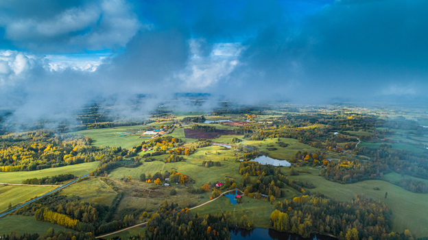 Aerial view of the rural landscape featuring lush green fields patches of autumn colored forests small lakes and dramatic clouds under a bright sky Latvia. Aerial view of the rural landscape featuring lush green fields patches of autumn colored forests small lakes and dramatic clouds under a bright sky Latvia.