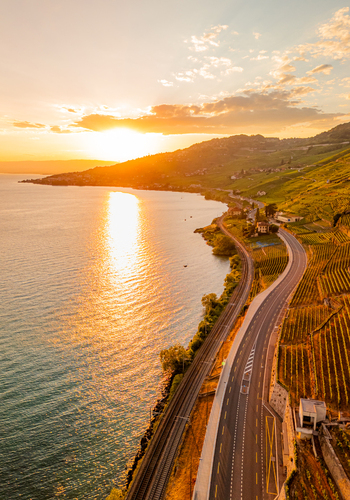 Aerial view of the terraced vineyards of Lavaux along the shores of Lake Geneva at sunset with a road and railway tracks in Bourg en Lavaux Vaud Switzerland. Aerial view of the terraced vineyards of Lavaux along the shores of Lake Geneva at sunset with a road and railway tracks in Bourg en Lavaux Vaud Switzerland.
