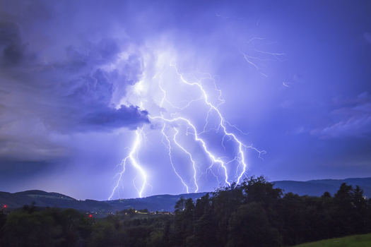 View of powerful lightning bolts striking from a dark stormy thunderstorm sky over the rolling hills and silhouettes of trees Graz Styria Austria.