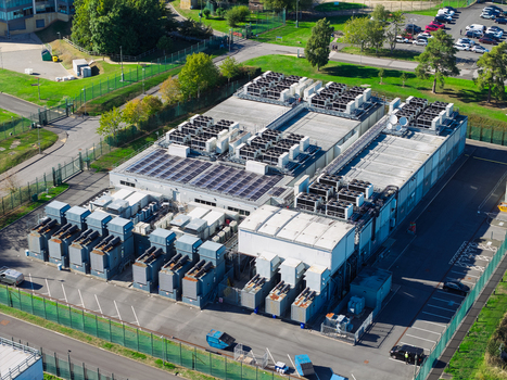 Aerial view of a large data center facility featuring extensive rooftop HVAC cooling units and solar panels surrounded by a fenced perimeter and parking area in the United Kingdom.