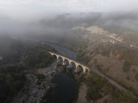 Aerial view of a stone railway bridge spanning a river in a valley surrounded by dense forests and rolling hills partially covered in morning fog in Balabanovo Kardzhali Province Bulgaria. Aerial view of a stone railway bridge spanning a river in a valley surrounded by dense forests and rolling hills partially covered in morning fog in Balabanovo Kardzhali Province Bulgaria.