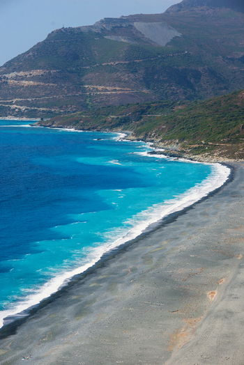 Aerial view of Plage de Nonza featuring dark grey pebbles and turquoise Mediterranean waters along the rugged mountains of Corse France.