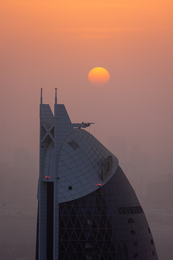 View of the modern architectural top of a skyscraper with the sun setting in a hazy orange sky above Dubai Dubai United Arab Emirates. View of the modern architectural top of a skyscraper with the sun setting in a hazy orange sky above Dubai Dubai United Arab Emirates.