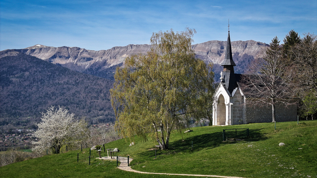 Aerial view of Chapelle Notre Dame de Riantmont on a grassy hill with mountains in the background in Vesancy Auvergne Rhone Alpes France. Aerial view of Chapelle Notre Dame de Riantmont on a grassy hill with mountains in the background in Vesancy Auvergne Rhone Alpes France.