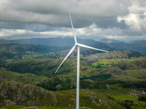 Aerial view of a wind turbine at Makaknuten Vindkraftverk set against a backdrop of rolling green hills rocky terrain and a cloudy sky in Bjerkreim Municipality Rogaland Norway. Aerial view of a wind turbine at Makaknuten Vindkraftverk set against a backdrop of rolling green hills rocky terrain and a cloudy sky in Bjerkreim Municipality Rogaland Norway.