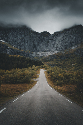 View of the dramatic Batman wall mountain face rising above a winding asphalt road surrounded by autumn foliage and moody clouds in Ramberg Nordland Norway. View of the dramatic Batman wall mountain face rising above a winding asphalt road surrounded by autumn foliage and moody clouds in Ramberg Nordland Norway.
