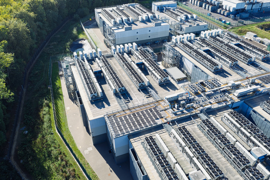 Aerial view of Data Center with rows of cooling units and industrial equipment on the roof surrounded by trees and a perimeter fence in the United Kingdom.